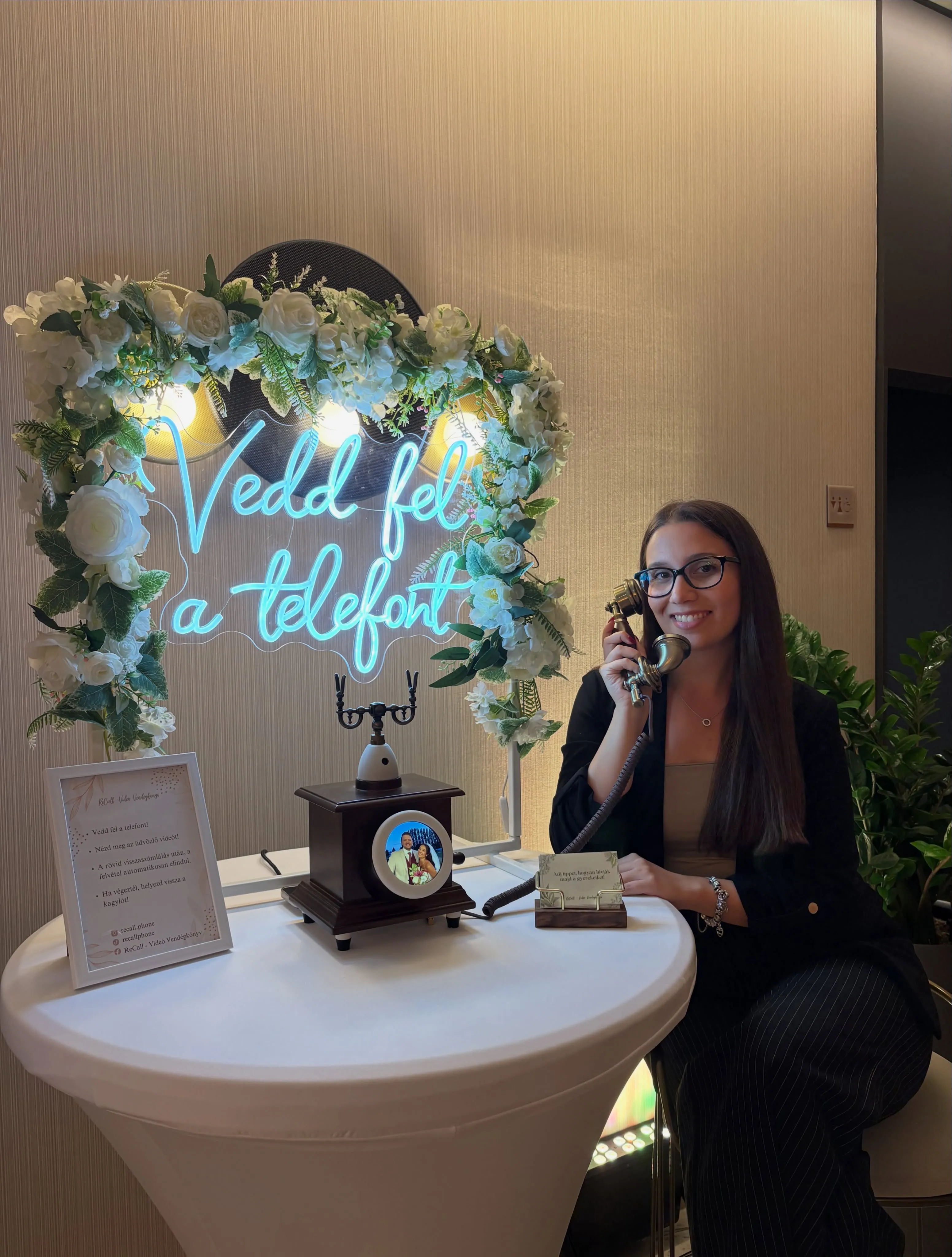 Szemüveges, blézeres vendég mosolyogva telefonál a ReCall készüléken, mellette a fehér virágíves „Vedd fel a telefont” neonfelirat